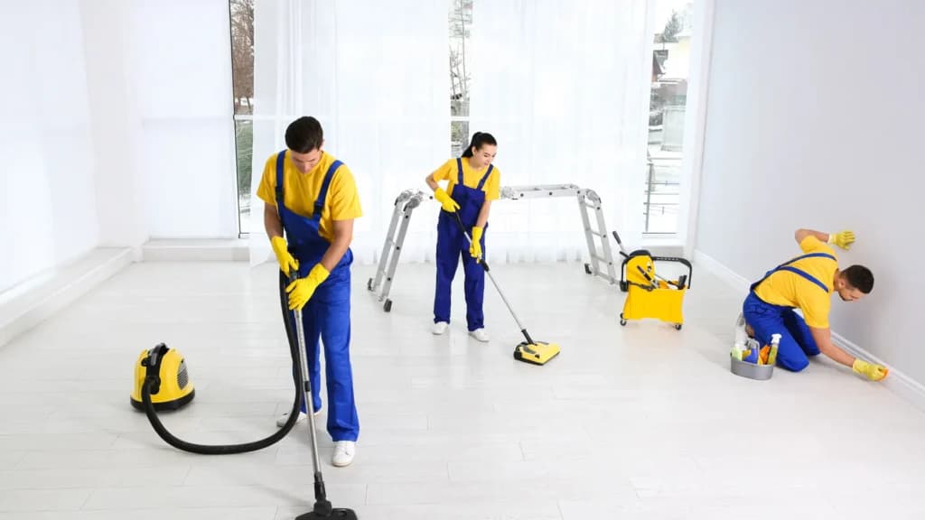 Three professional cleaners in blue and yellow uniforms vacuuming, mopping, and scrubbing a bright room.