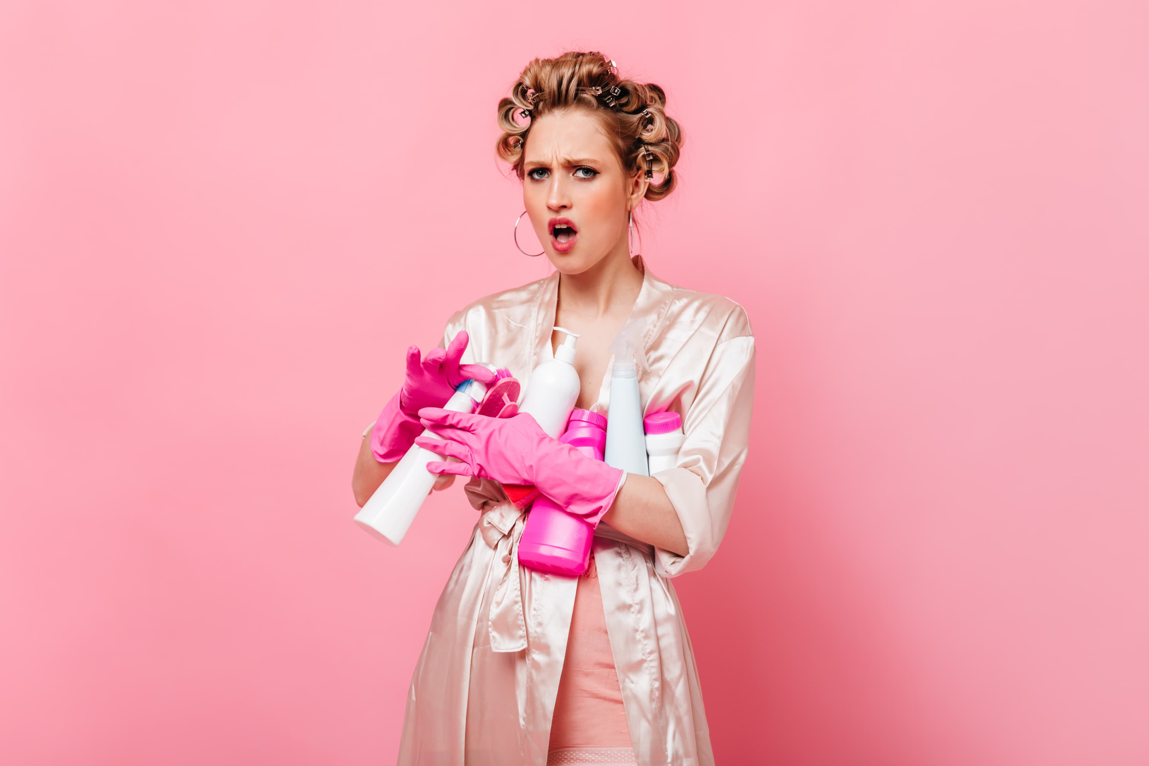 Surprised woman in hair curlers and robe clutching cleaning supplies against a pink background.
