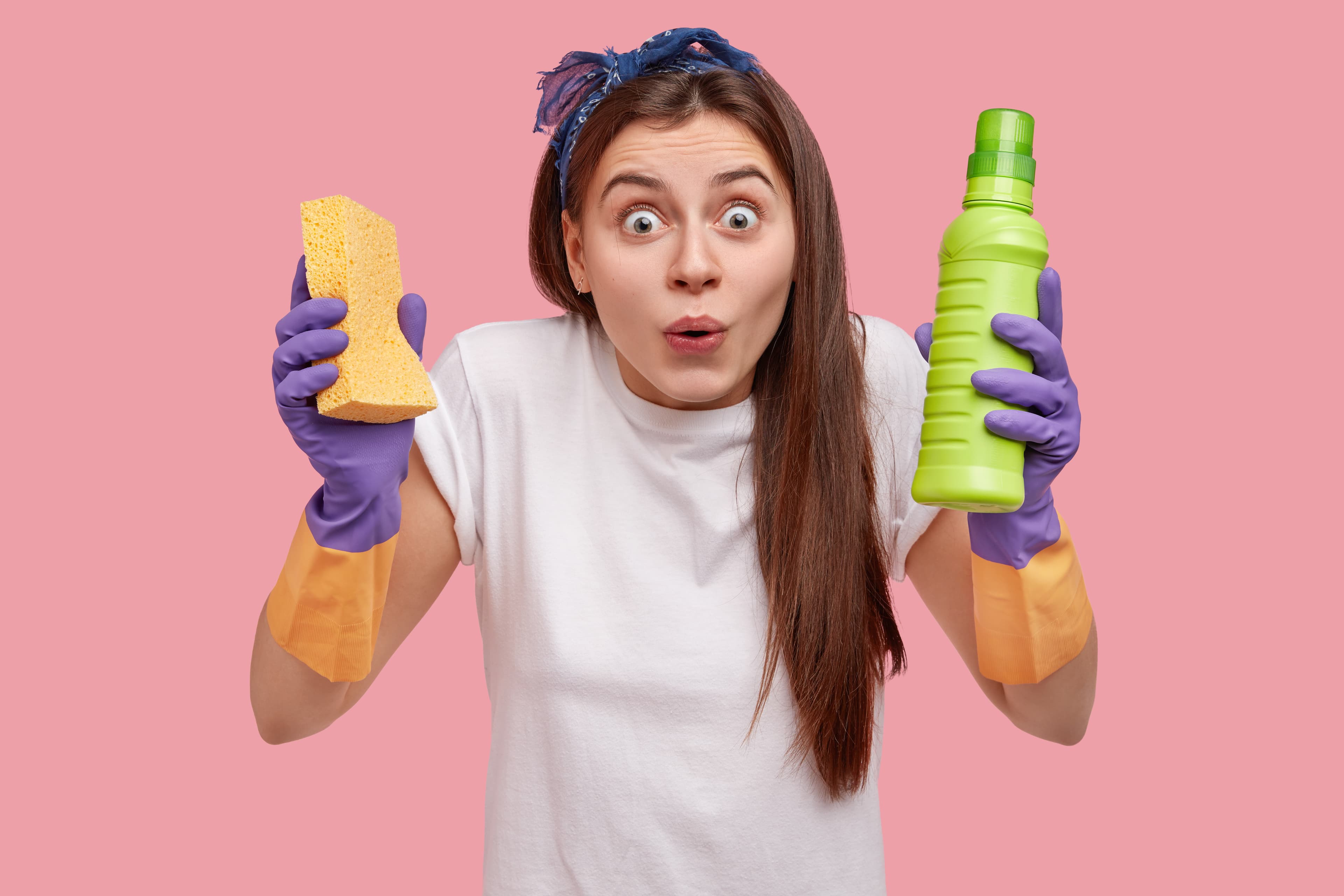 Surprised woman in cleaning gloves holds a sponge and green bottle against a pink background.