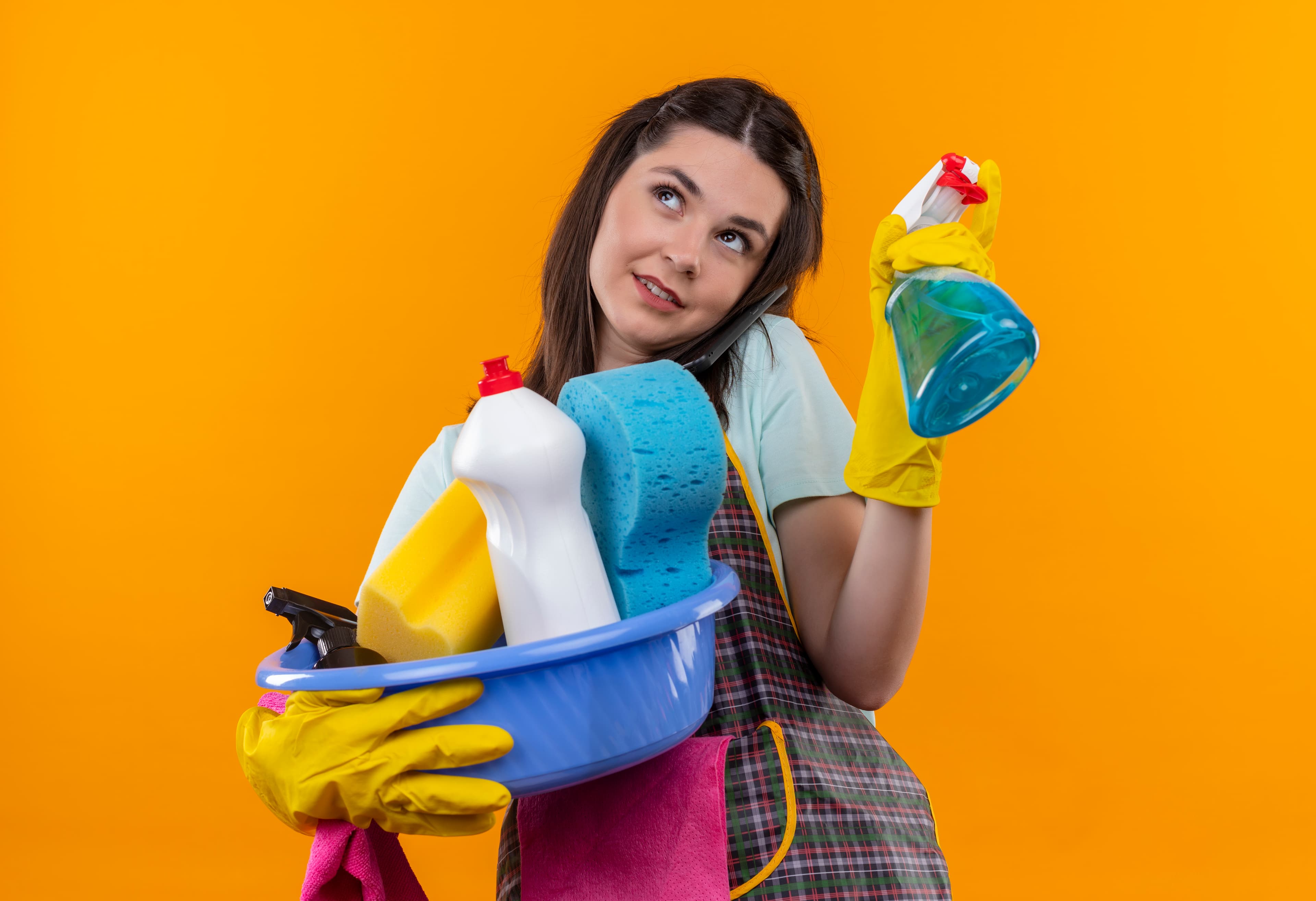 Woman in apron and yellow gloves holding cleaning supplies while talking on a phone.